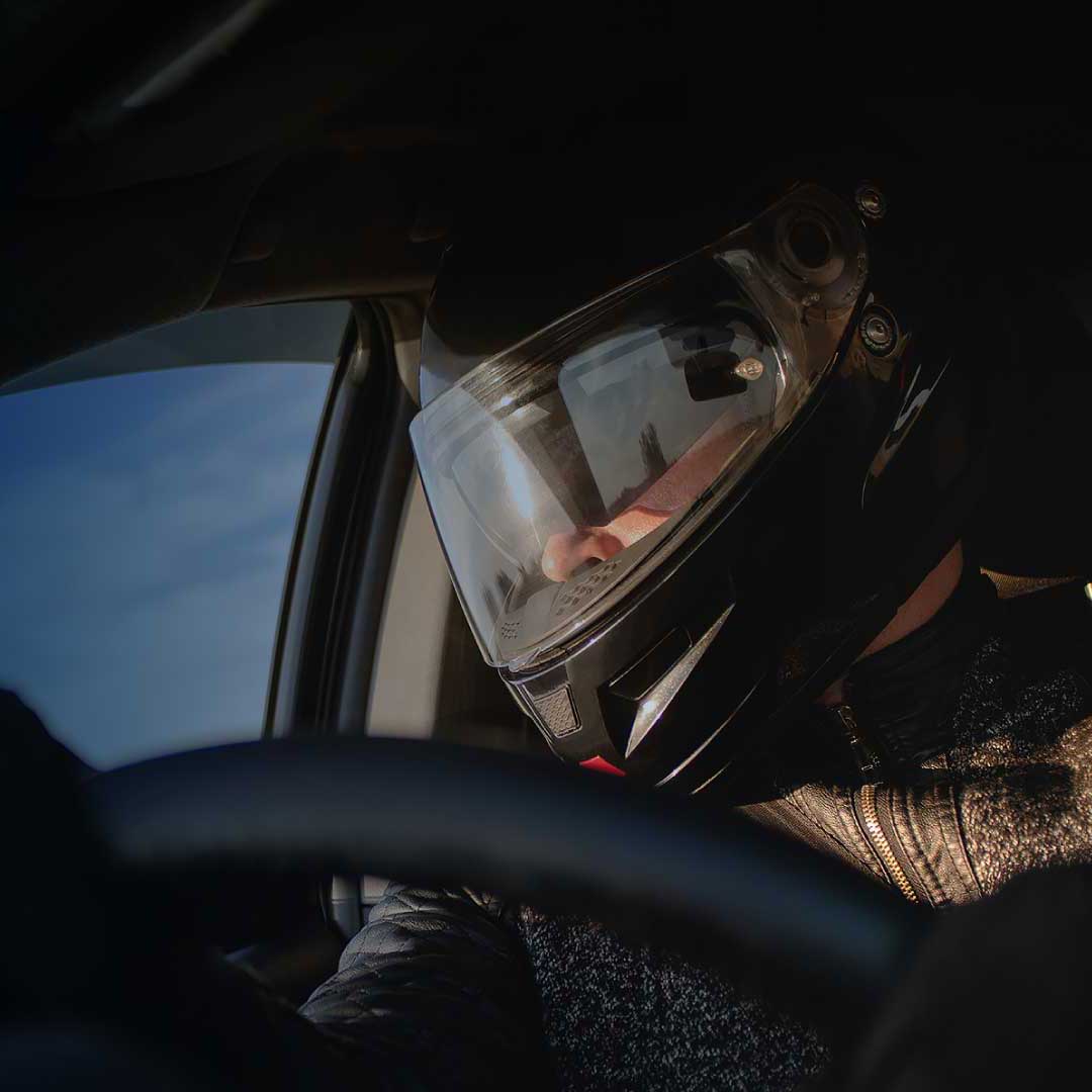 A race car driver grips the wheel, clad in a black helmet with a gleaming face shield.