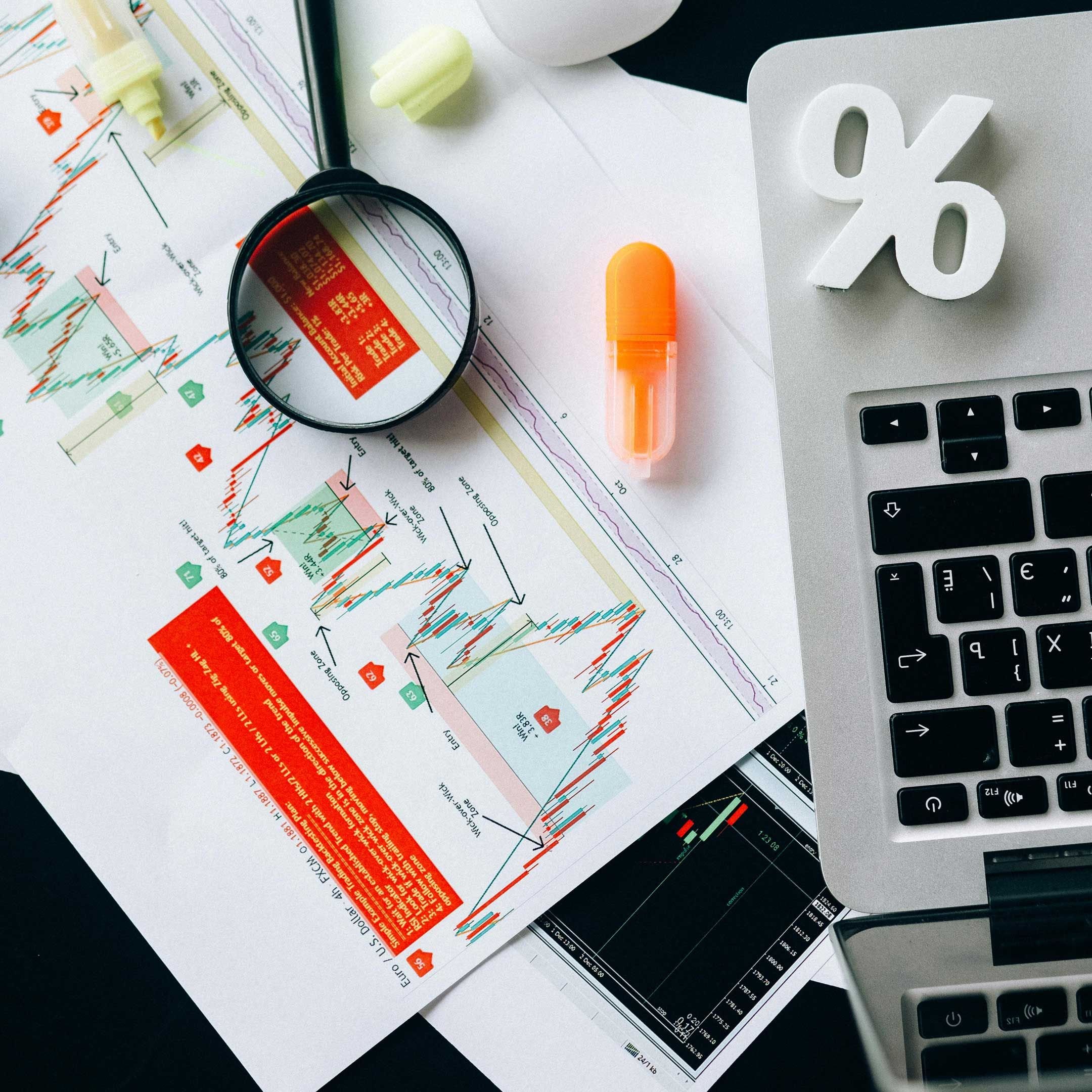 Image of items on a desk including financial paperwork, a magnifying glass, whiteout correction tape, a highlighter pen, a paperweight formed to look like a percent symbol, and a laptop